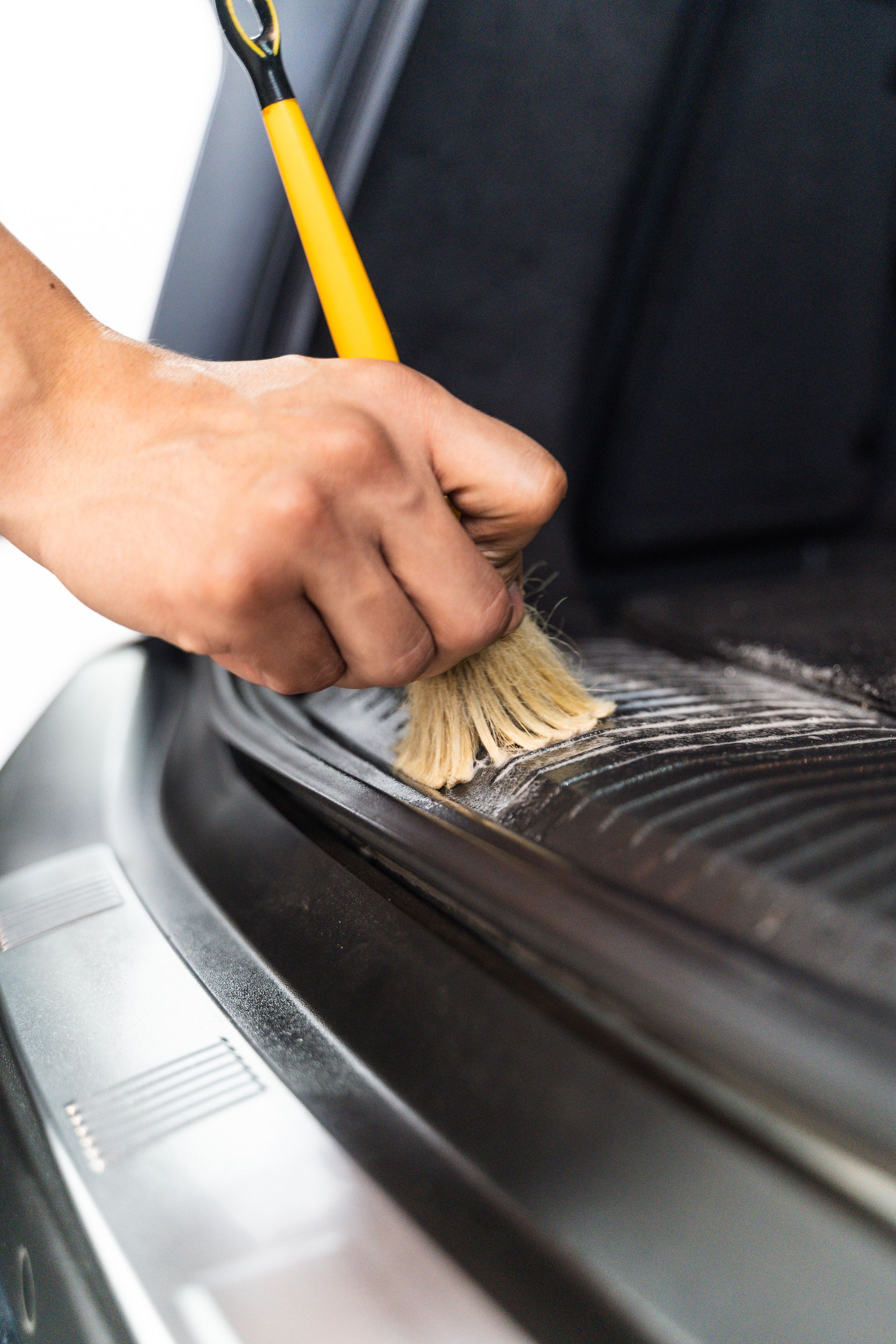 Person brushes dirt from a black rubber car mat with a yellow-handled brush.