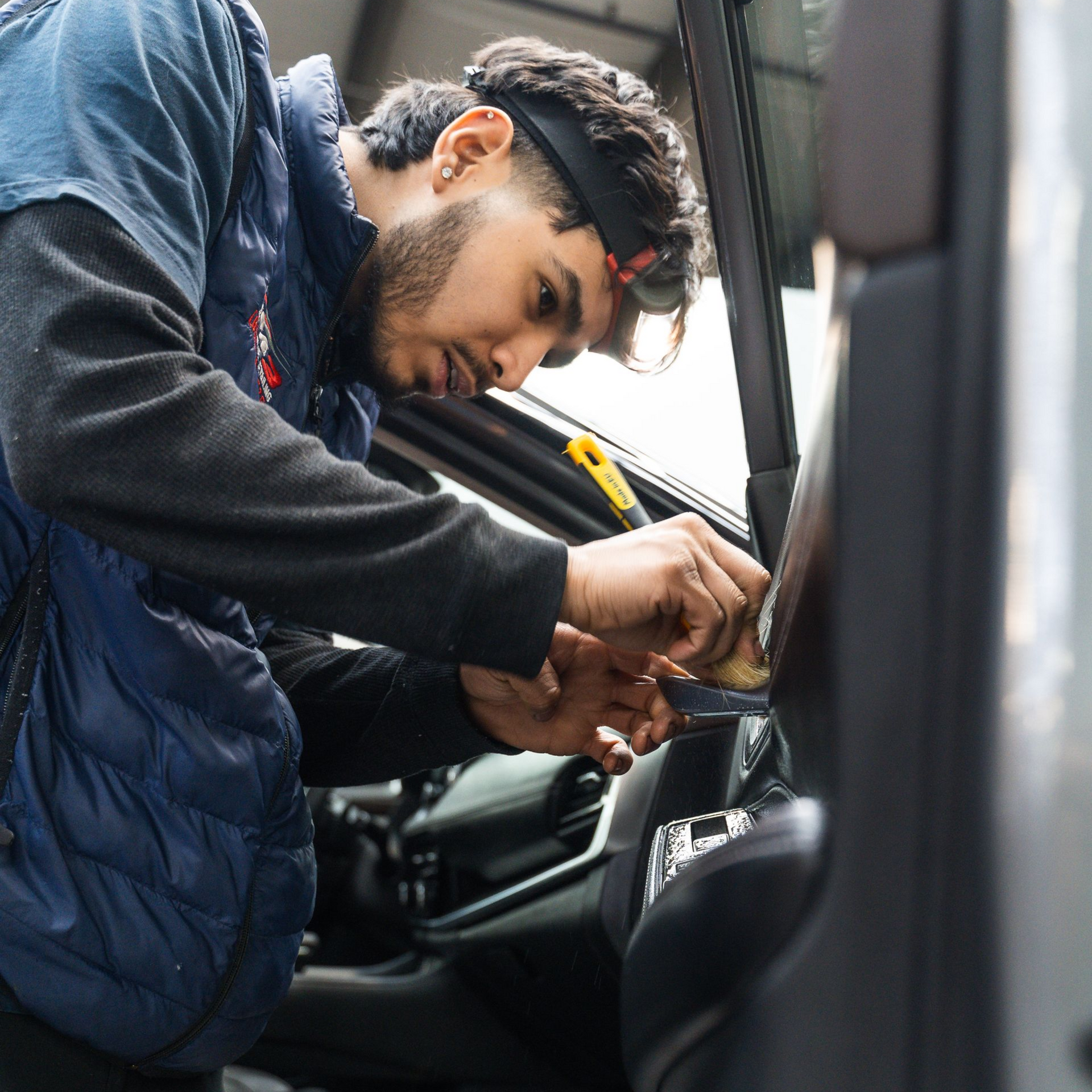 Man in blue vest works on car interior; using tools, focused.