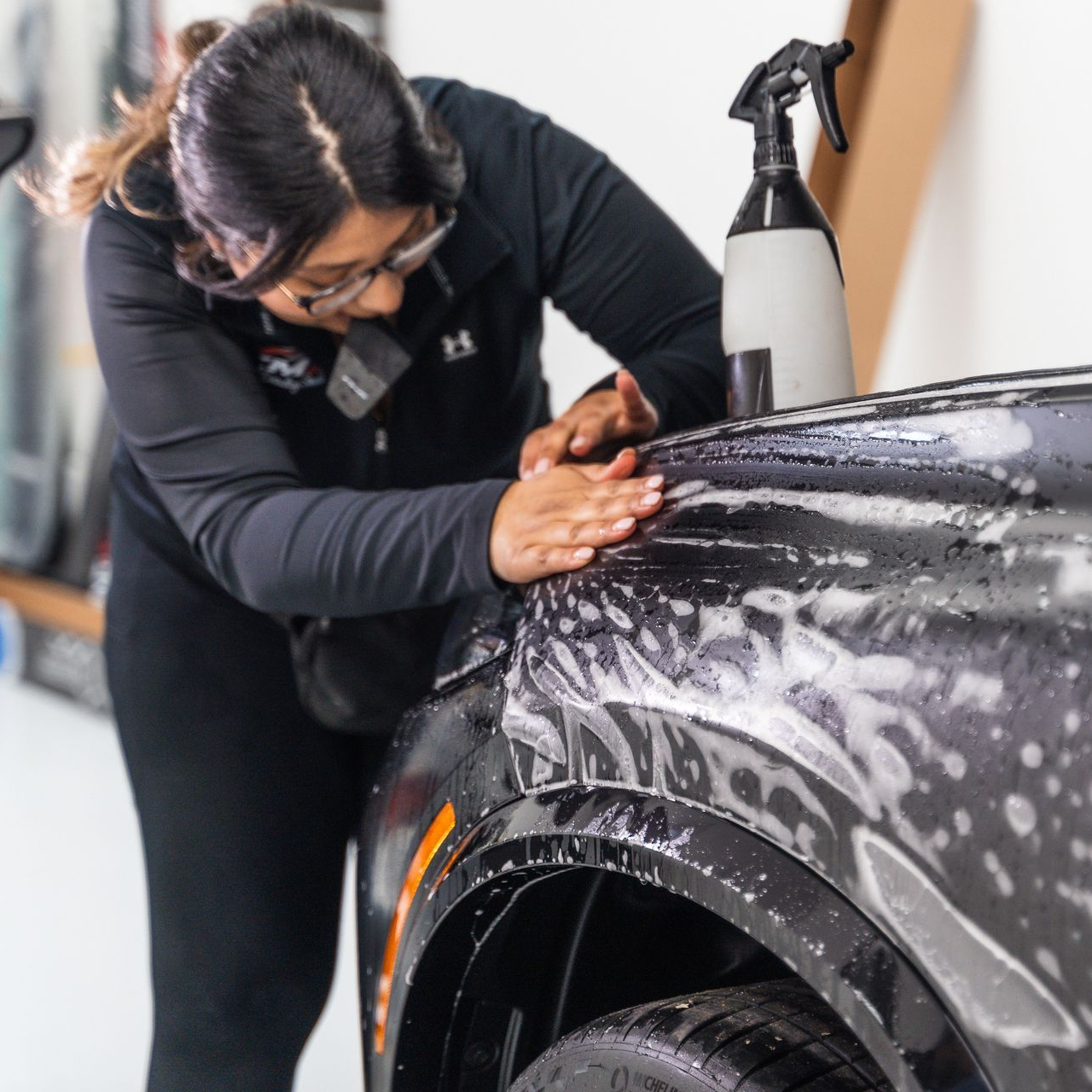 A person in a black jacket carefully applies a clear paint protection film to the front fender of a dark vehicle.
