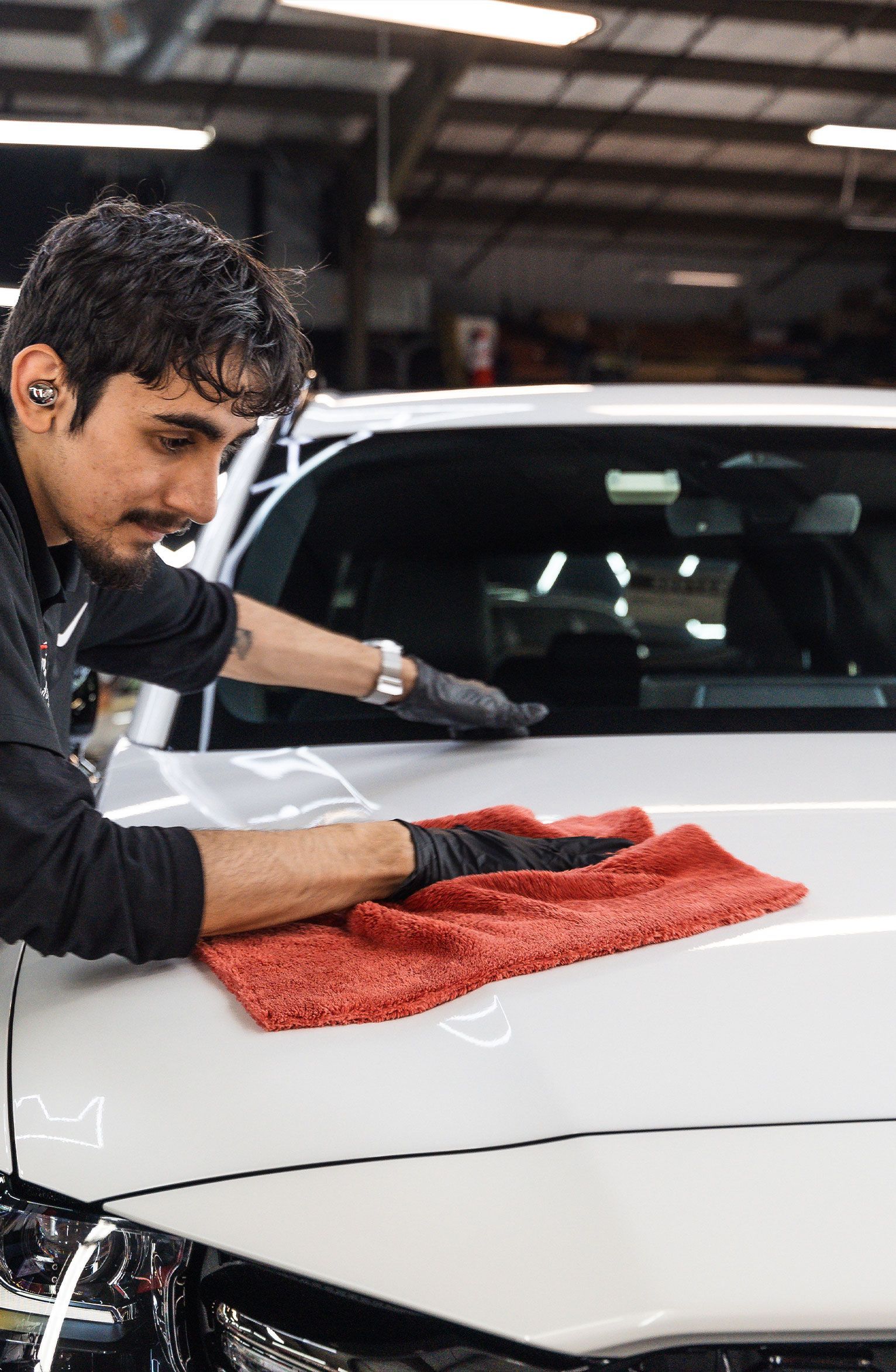 Person in black gloves wiping a white car with a red cloth in a garage.