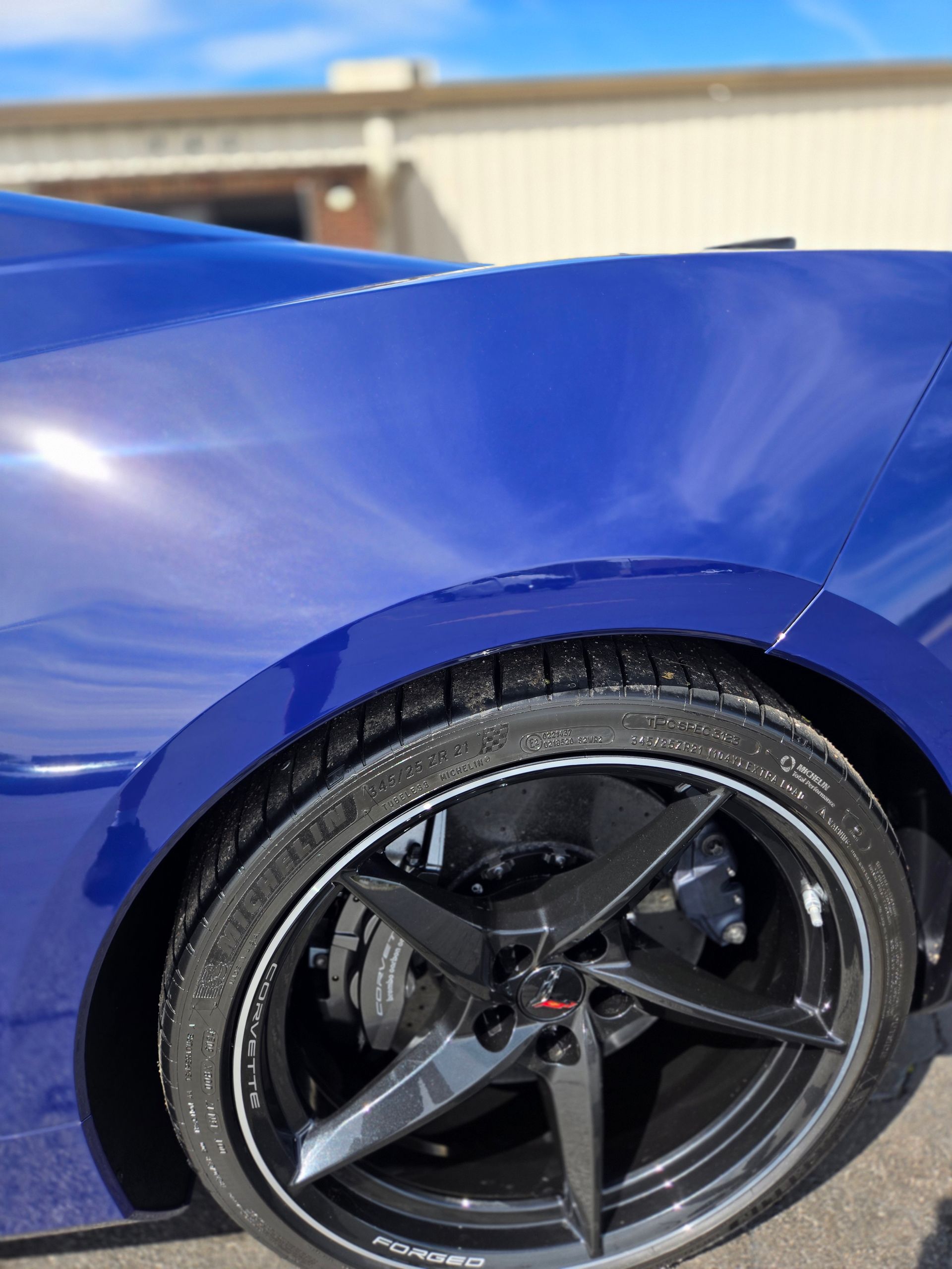 Blue sports car wheel well and tire, with a dark-colored rim, against a building.