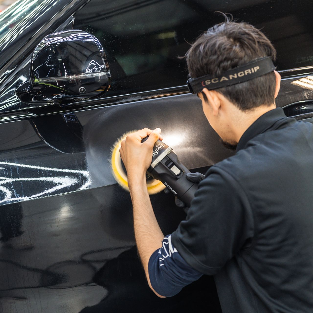 Person polishing black car with a rotary buffer, wearing a headlamp.