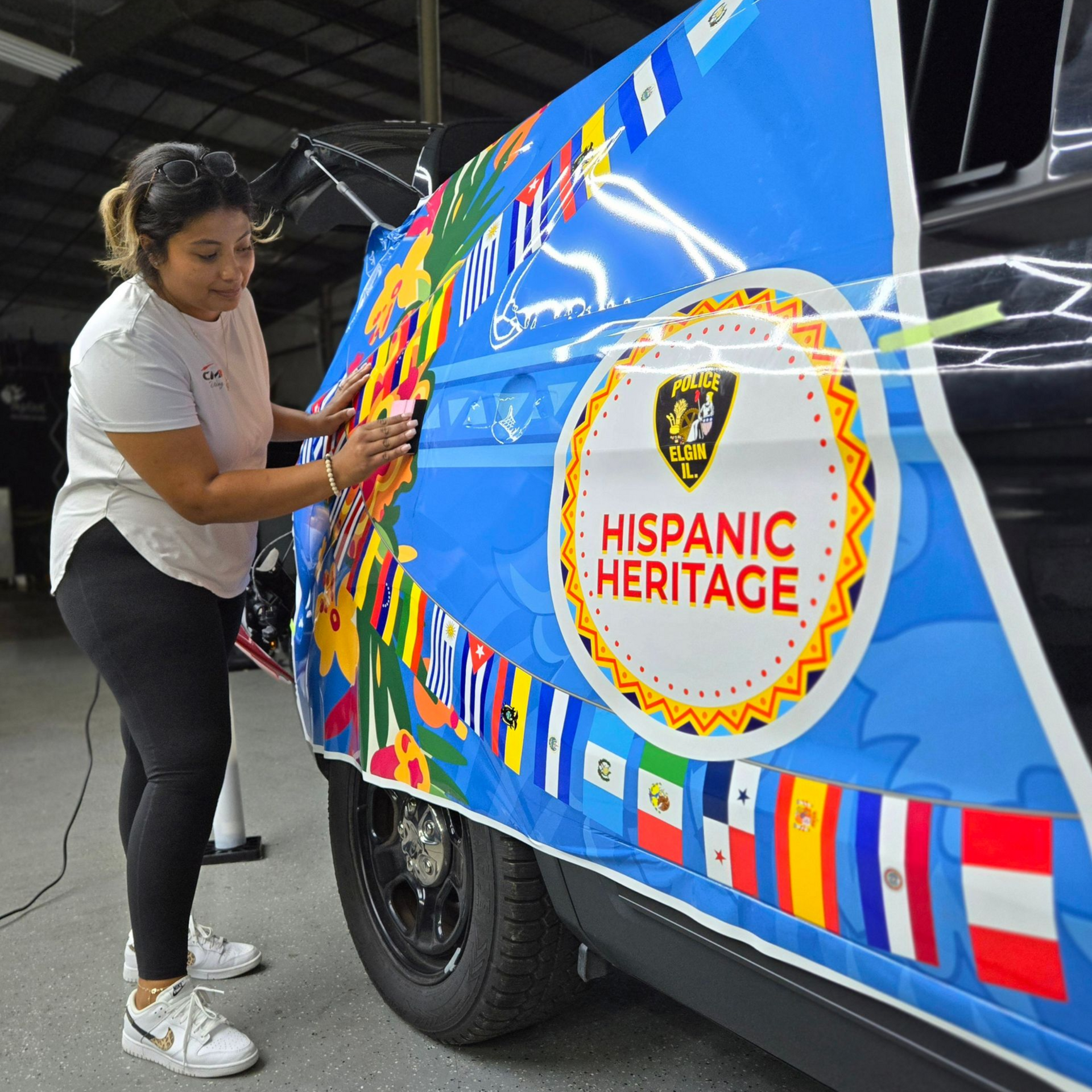 Two people wrapping a car's rear bumper with blue film inside a shop; one person kneels, applying the film.