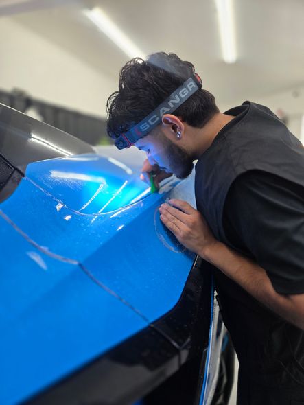 Man installing blue wrap on a car, illuminated by headlamp, inside a shop.