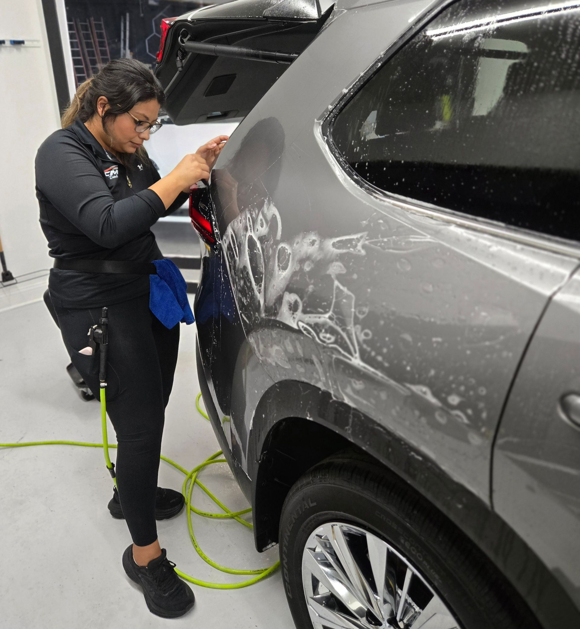Woman applying film to a gray car's rear quarter panel in a shop.
