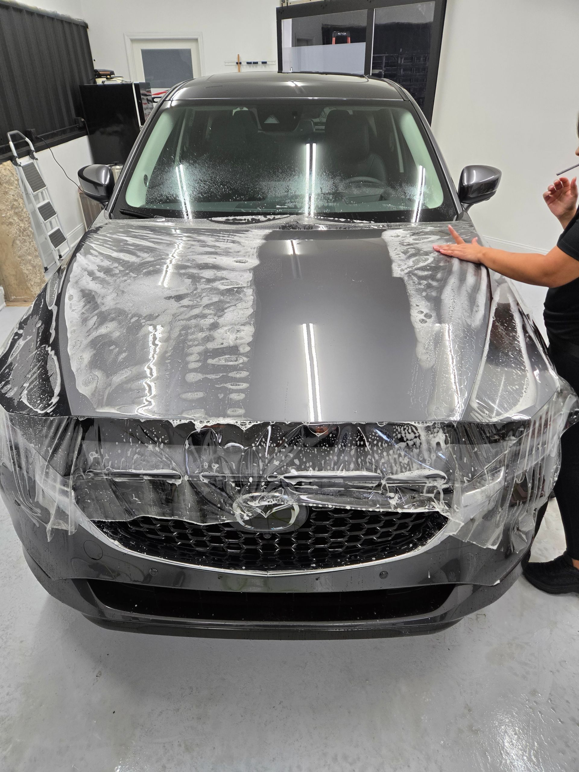 A person applying a protective film to the hood of a dark gray car in a shop.