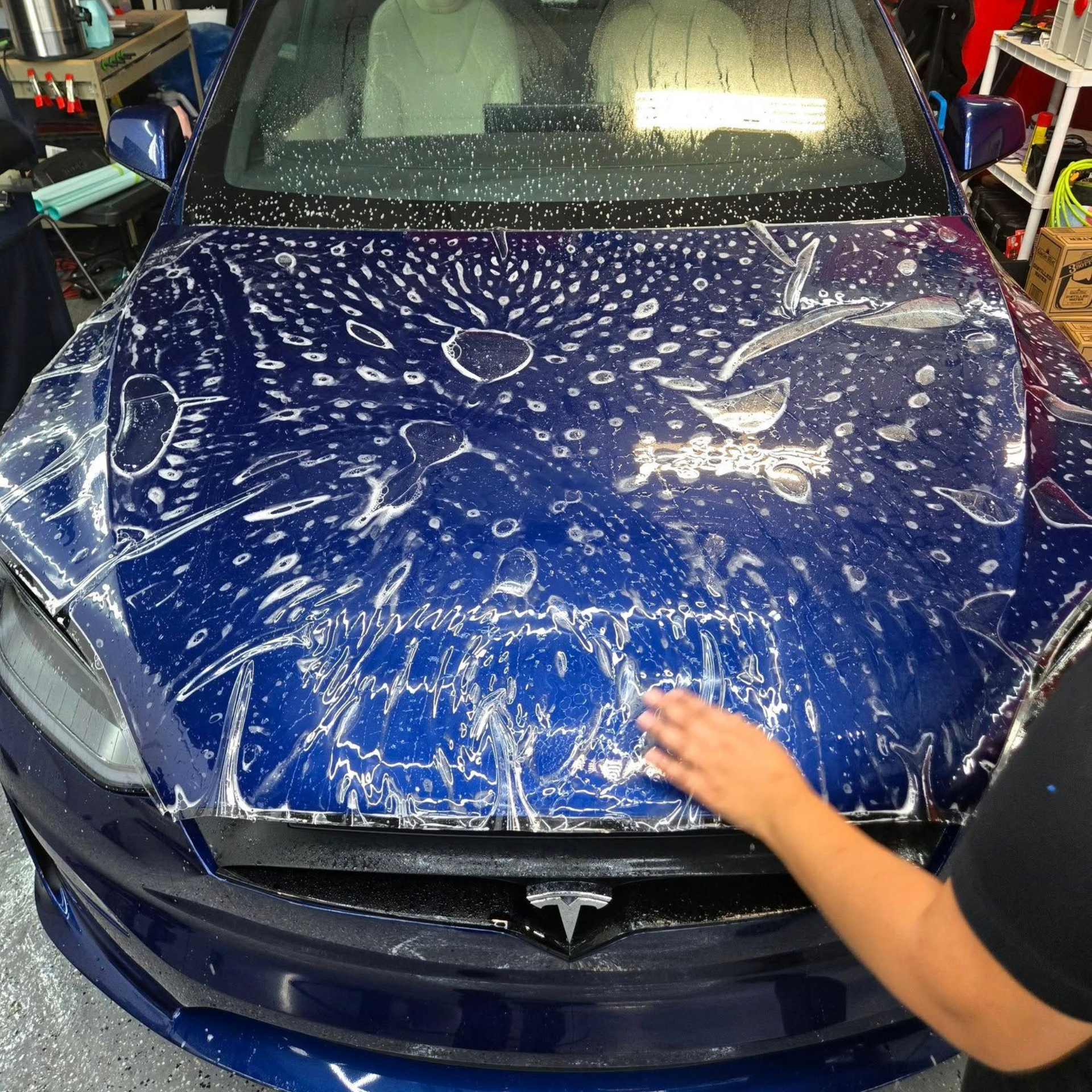 Person washes a blue car covered in soap, squatting near a black tire, in a garage.