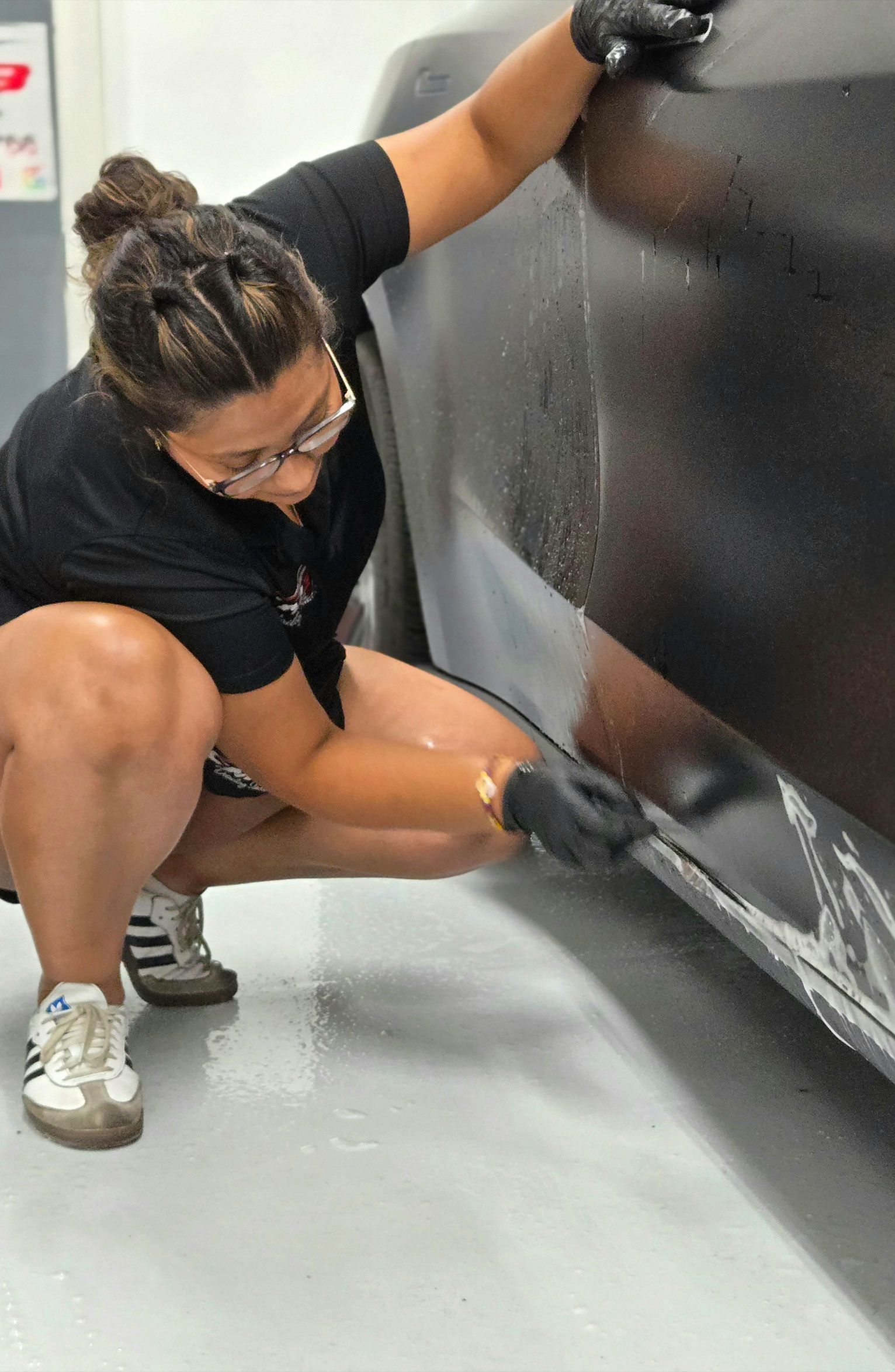 Person applying film to a car; indoors, squatting, black gloves, black shirt, Adidas sneakers, white film.