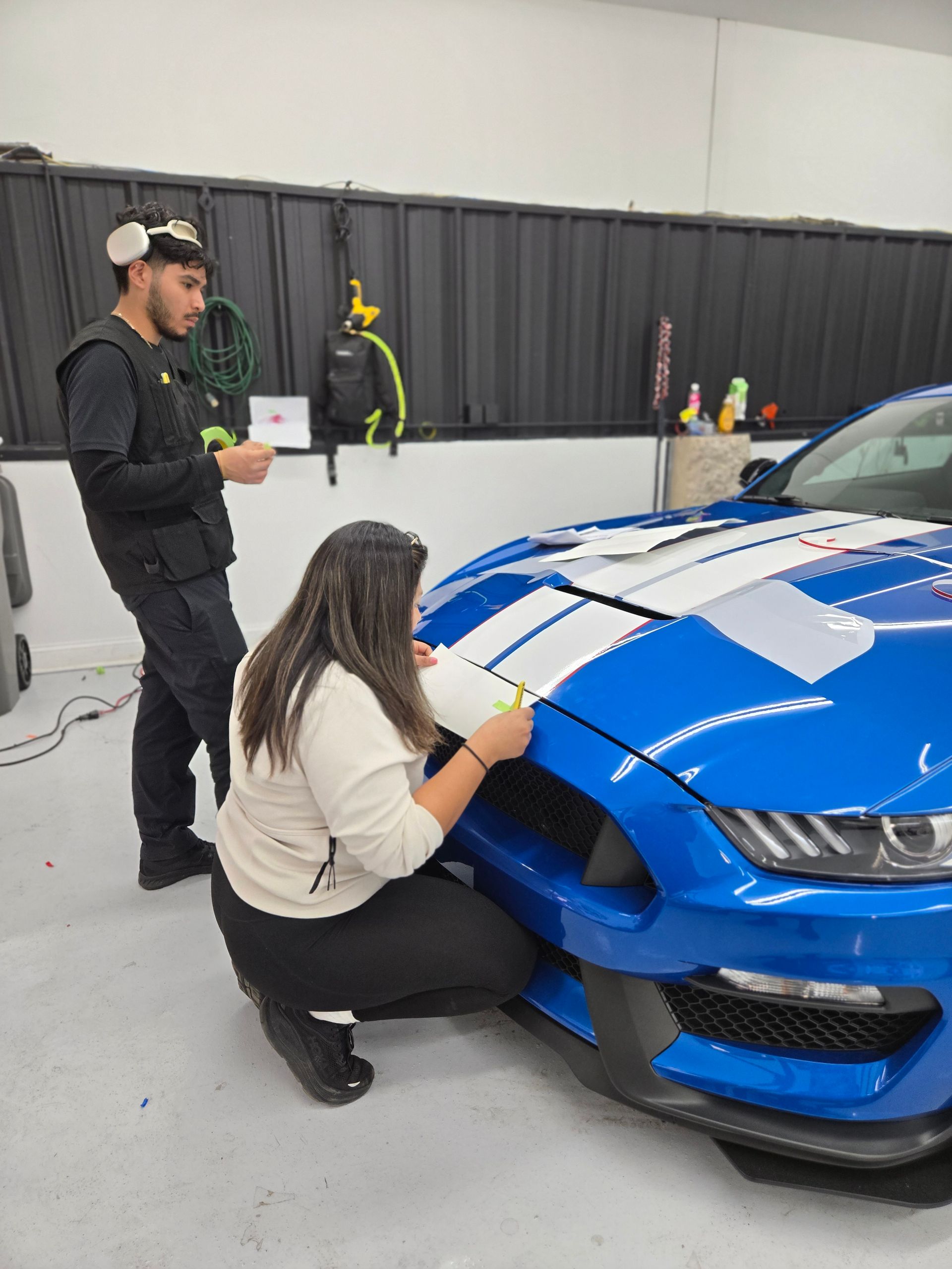 Two people applying white stripes to a blue Ford Mustang in a garage.