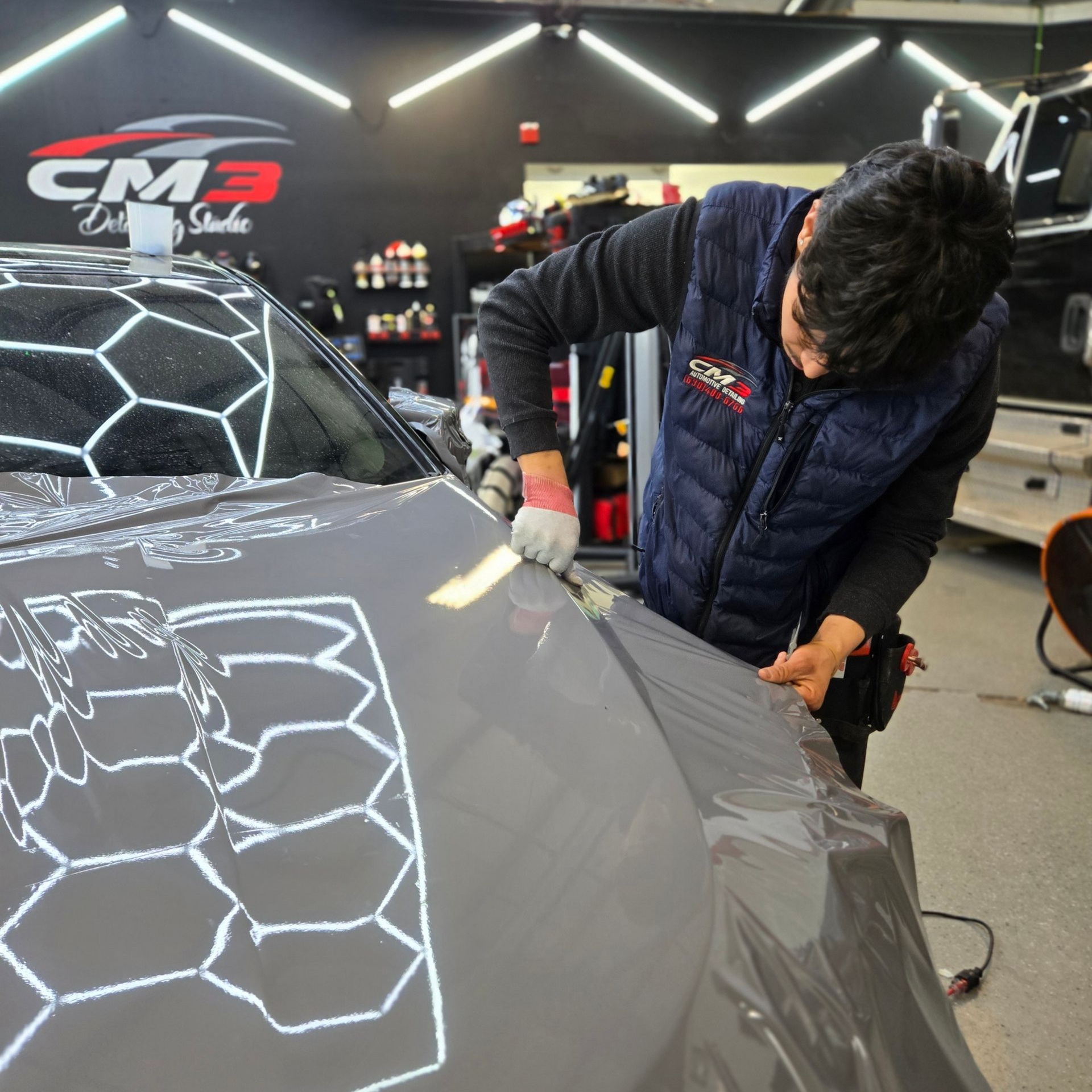 A person in a dark vest applies a grey protective film to a car hood in a studio with hexagonal overhead lighting.