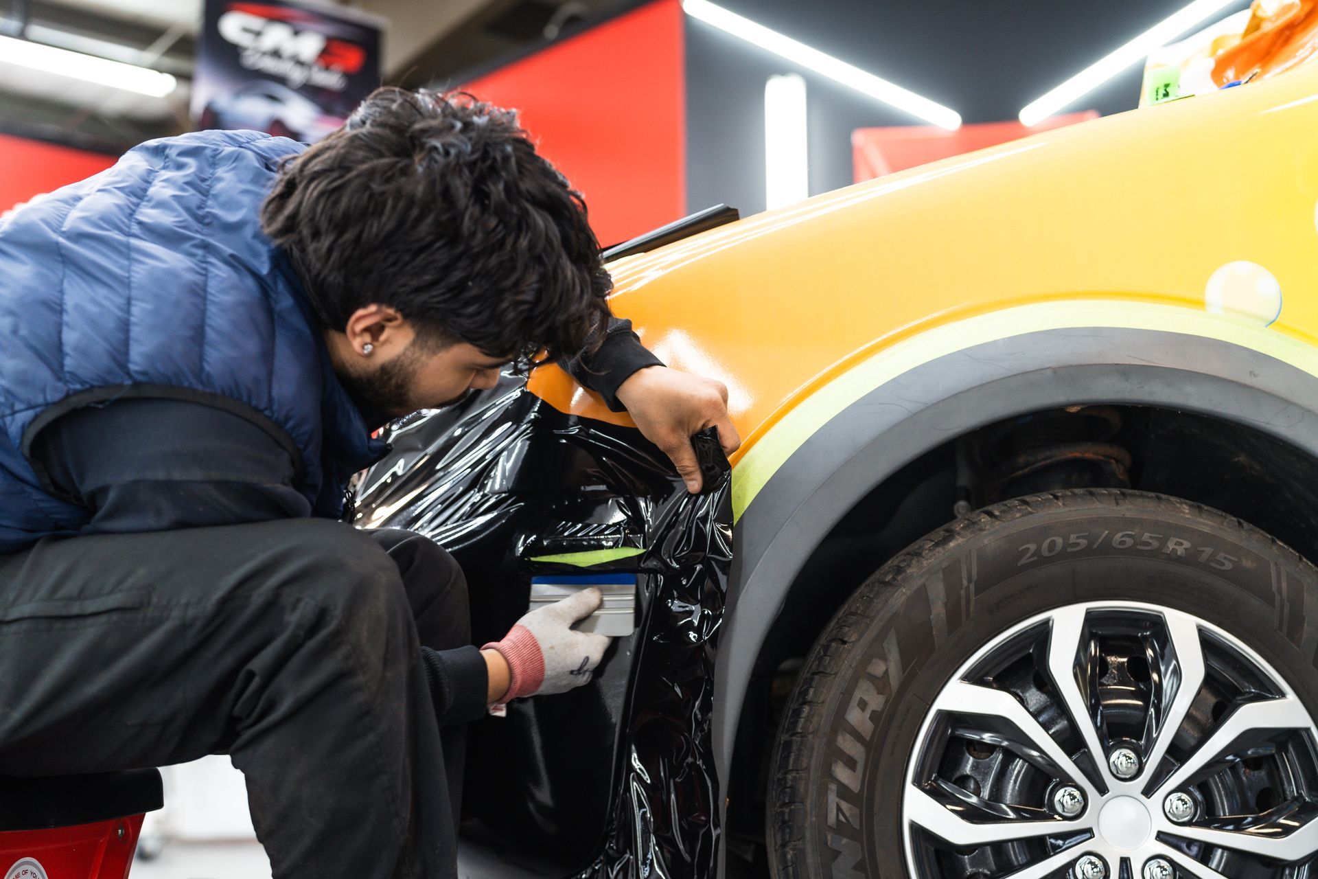 A person wraps a black film on a car fender in a workshop. The car is orange with black trim.