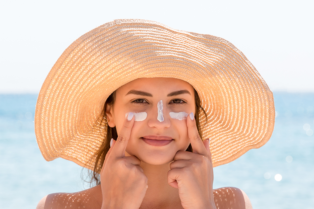 Eine Frau mit Strohhut trägt Sonnencreme auf ihr Gesicht auf.