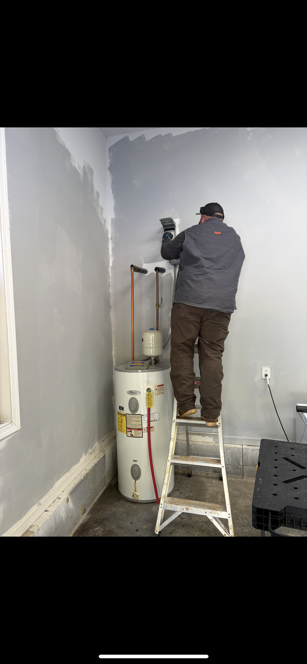 A person on a ladder is working on a wall near a water heater in a room.