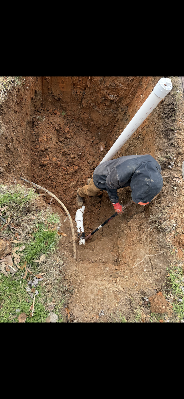 A person is working in a dug-out trench, installing piping in an outdoor setting.