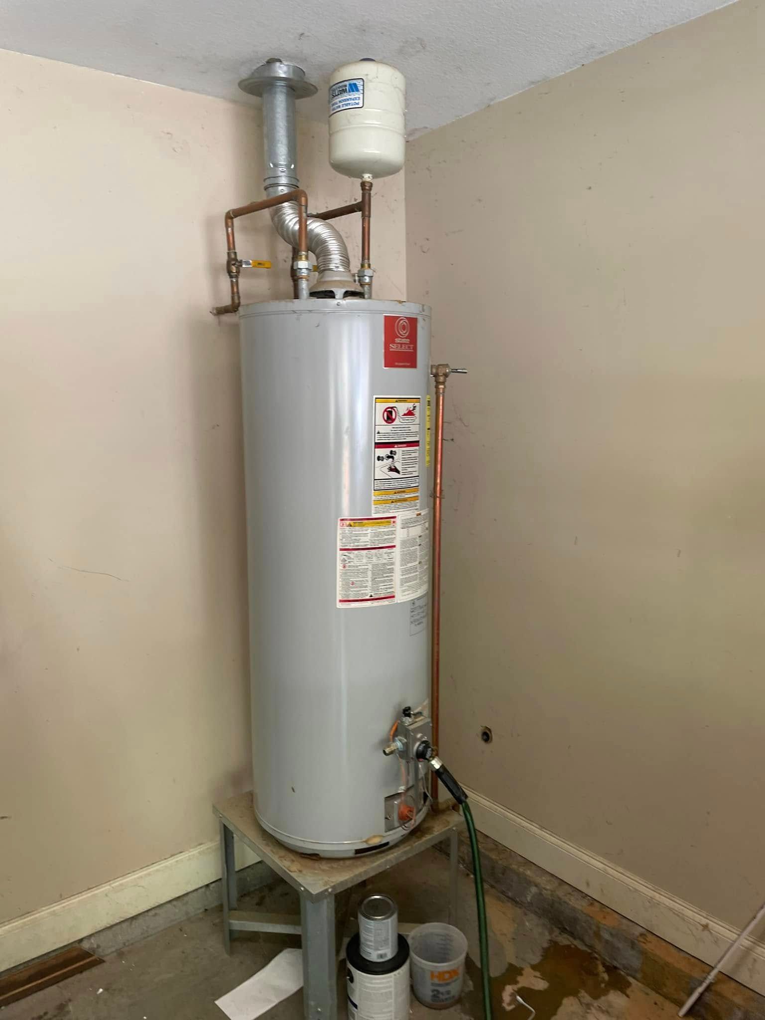 Water heater in a corner, with vent pipe and expansion tank. Gray and copper pipes against beige walls.