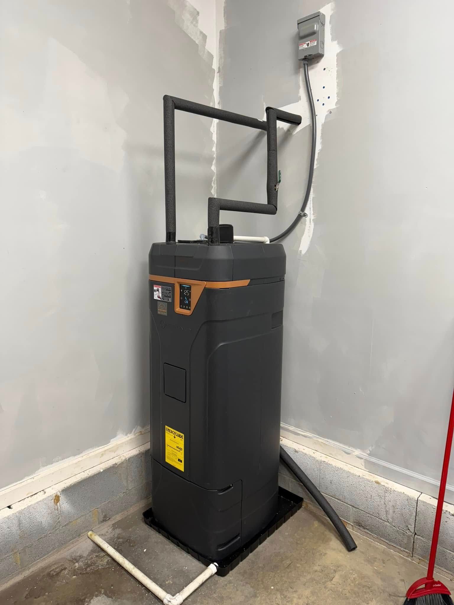 A black heat pump water heater in a garage corner, with black piping and a control panel.