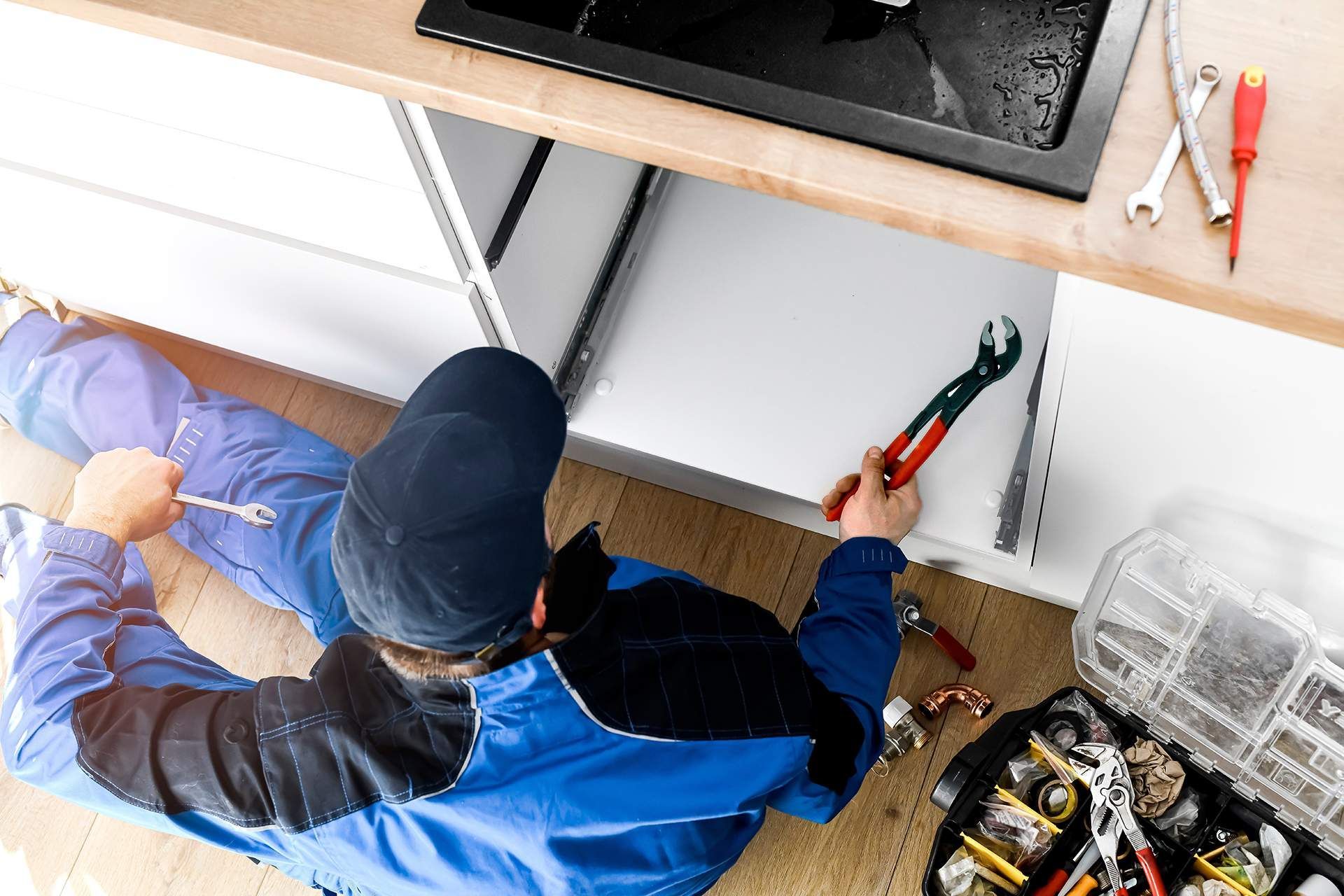 Plumber working under kitchen cabinet, using pliers. Toolbox and tools on the floor.