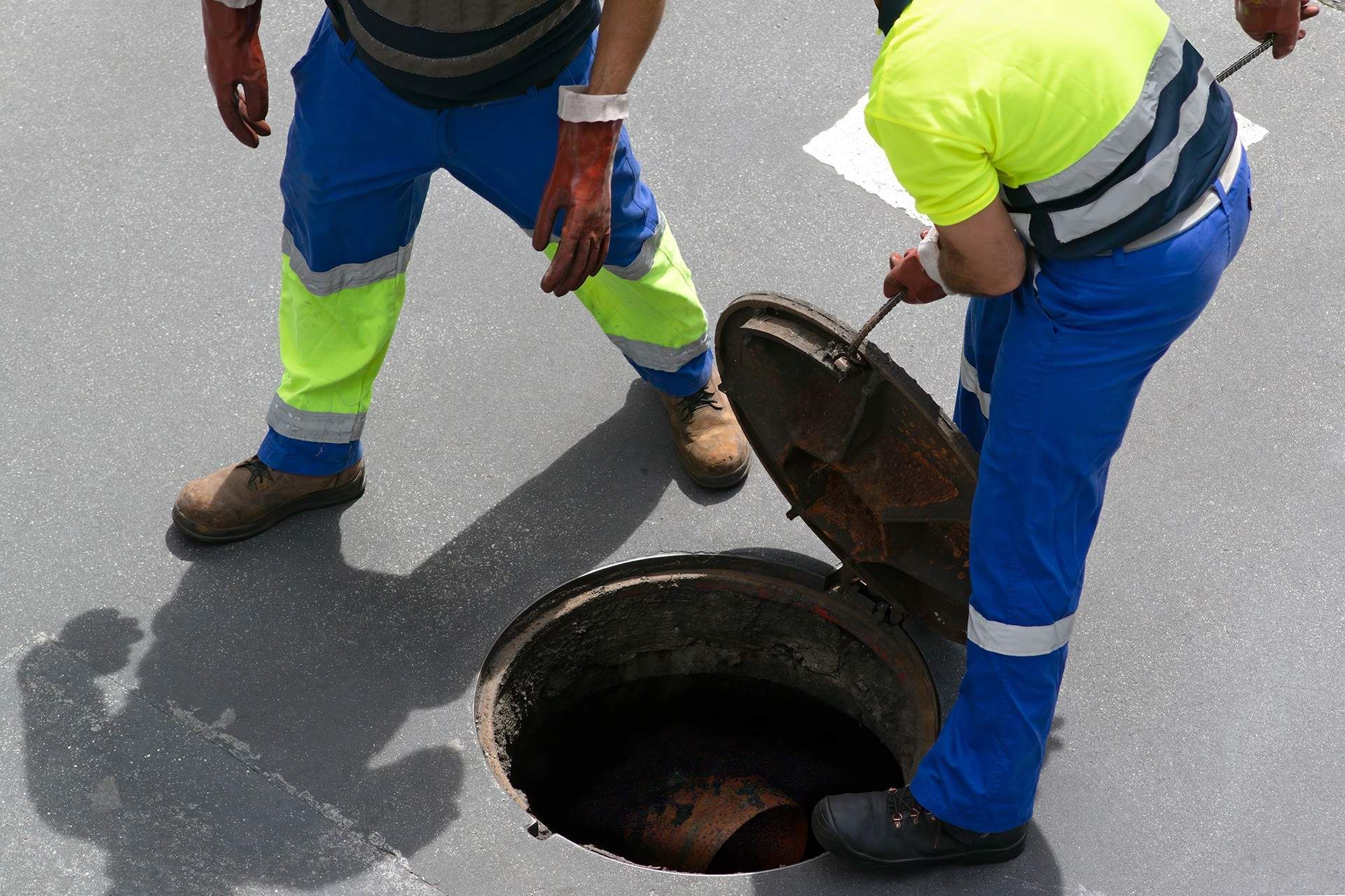 Two workers in blue and neon yellow safety gear open a manhole in the road.