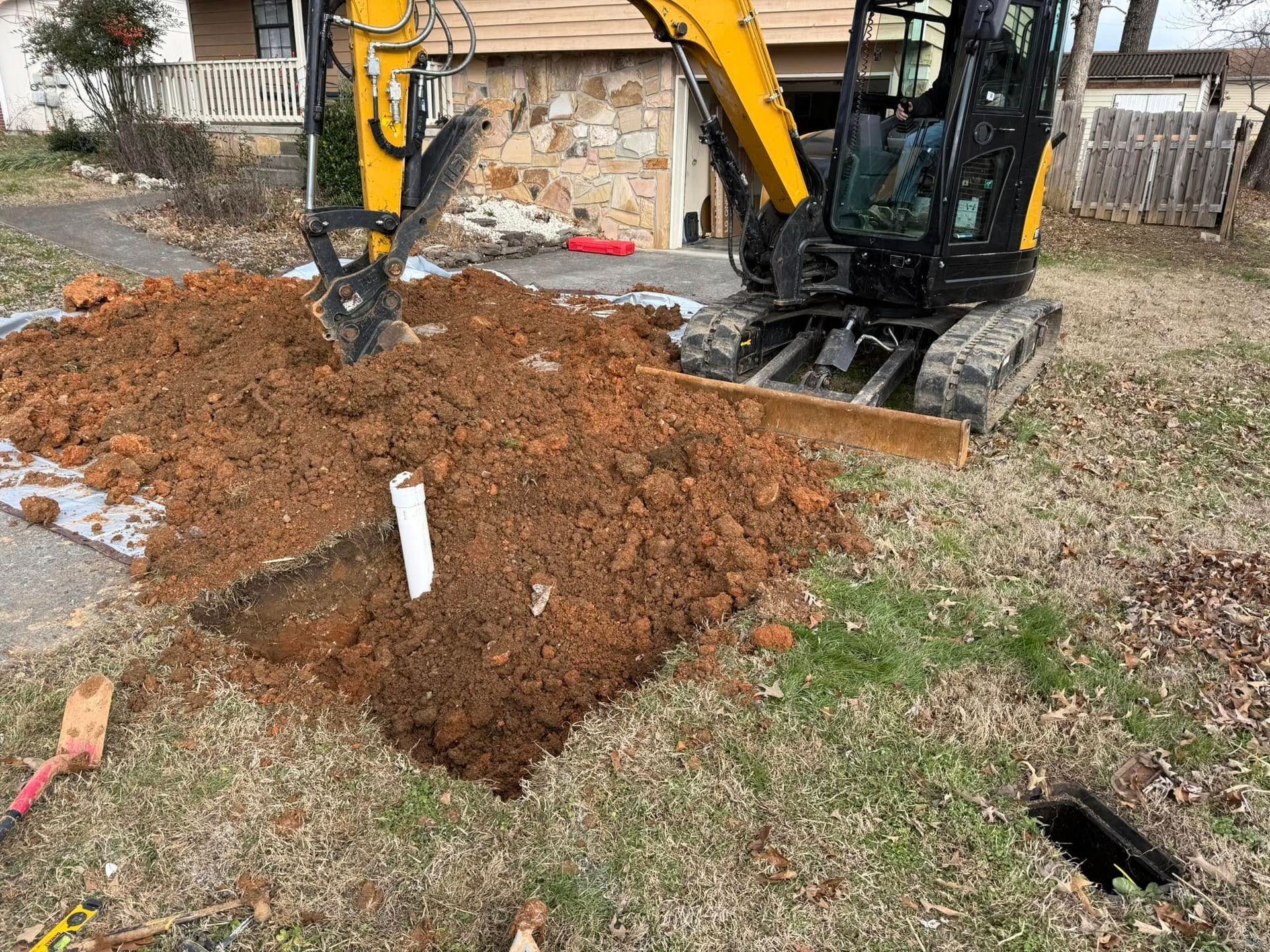 Yellow excavator digs a square hole in a grassy yard, exposing a white pipe. Brown dirt is piled up nearby.