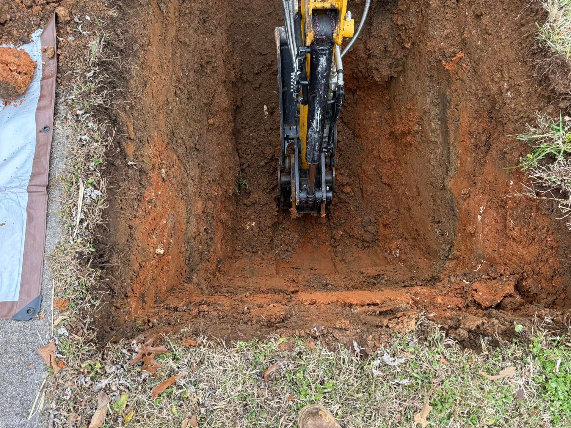 Mini excavator digging a square hole in reddish-brown soil, surrounded by grass.