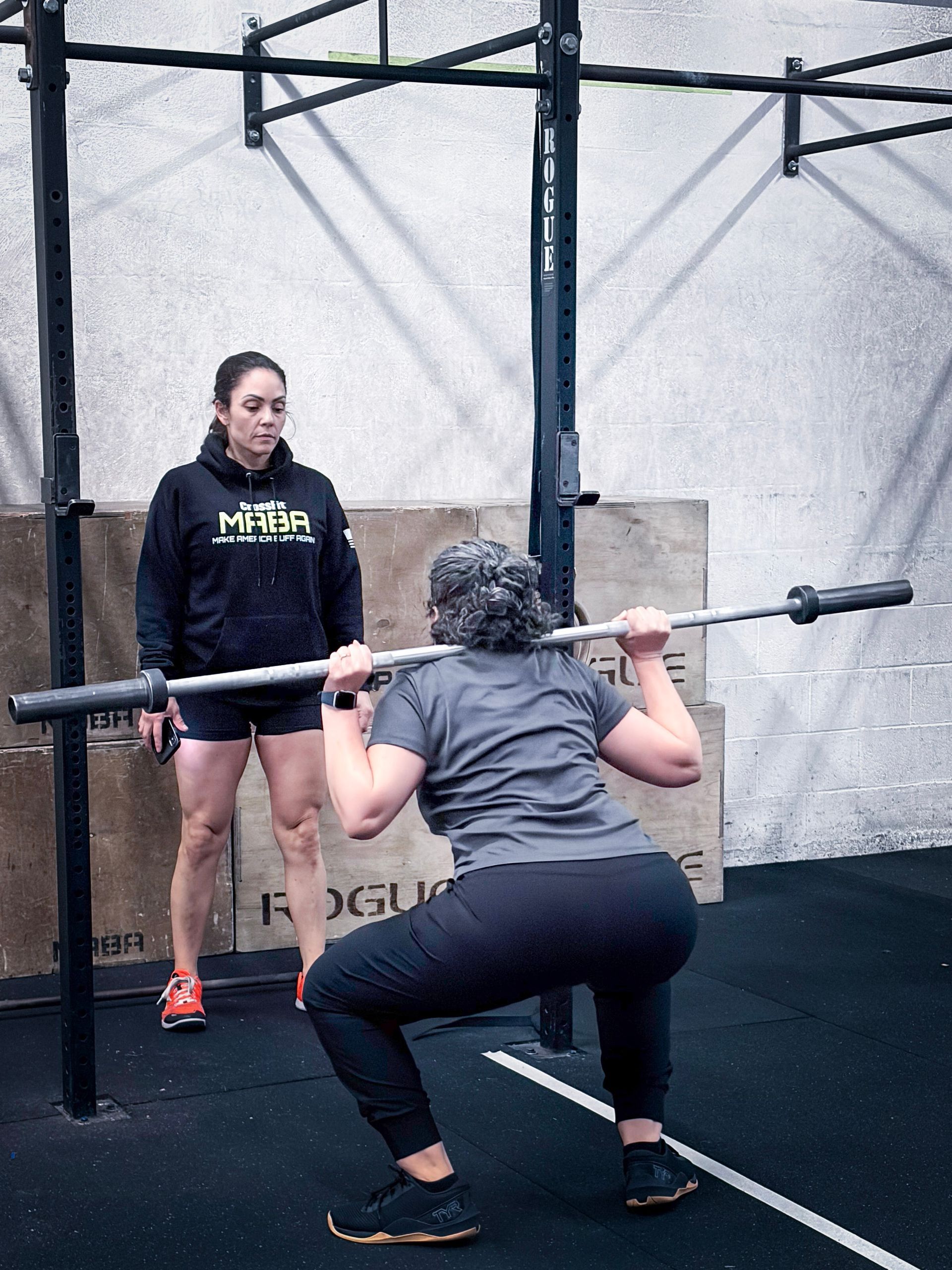 Woman doing a barbell squat with a trainer, inside a gym.