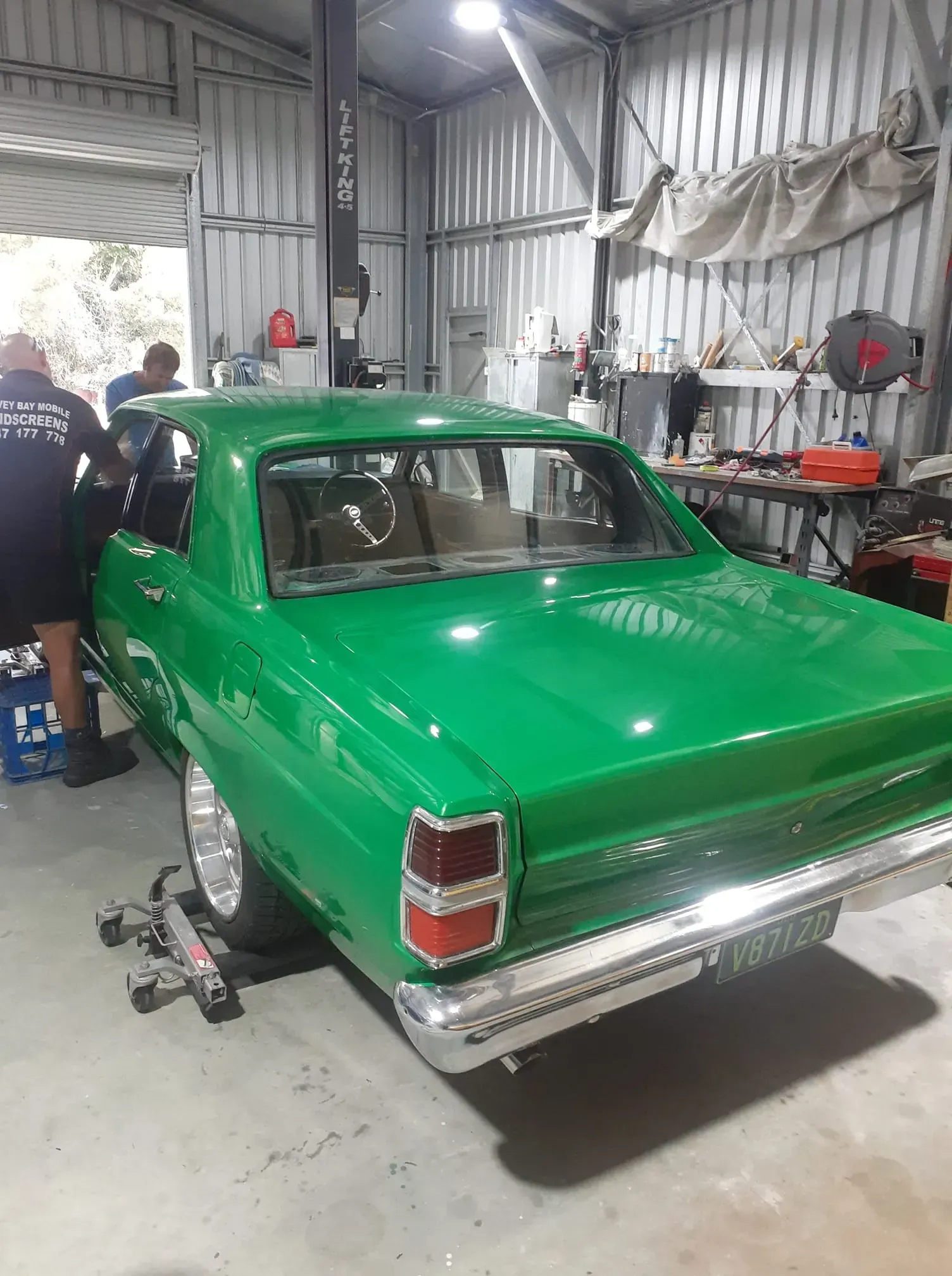 Green classic car in a workshop, under a car lift, with people working around it. — Hervey Bay Mobile Windscreens in Pialba, QLD