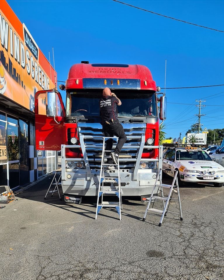 Man on ladder working on the front of a red truck in front of a building with a sign. — Hervey Bay Mobile Windscreens in Pialba, QLD