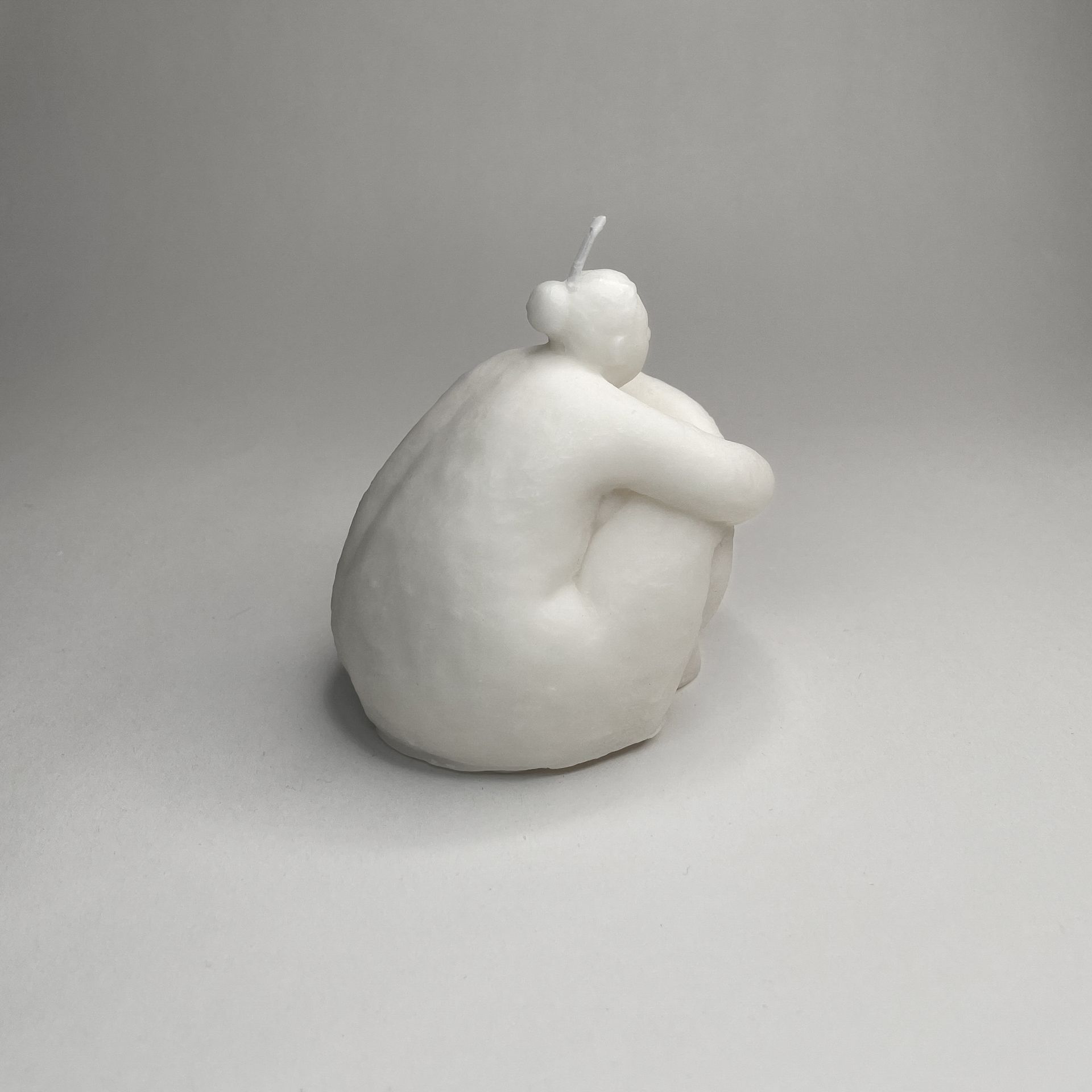 Een witte kaars in de vorm van een vrouw zit op een wit oppervlak.