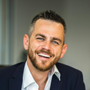 Man with short hair and beard smiles, wearing a navy blazer and white collared shirt.