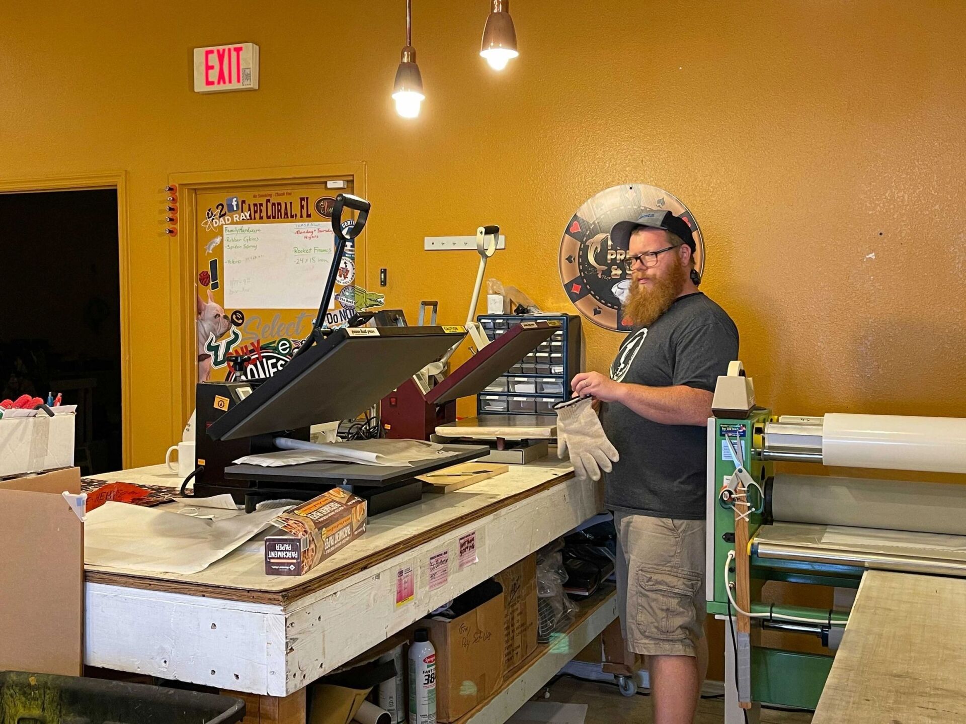 Man with beard prepares equipment in a print shop; heat press, work table, and printed materials are visible.