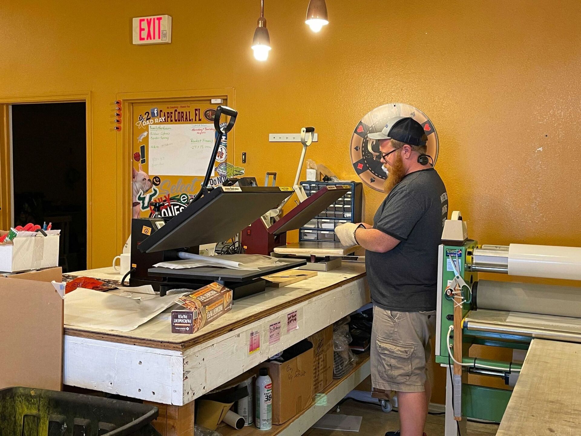 Man operating a heat press in a workshop. Yellow walls, white workbench, and an exit sign.