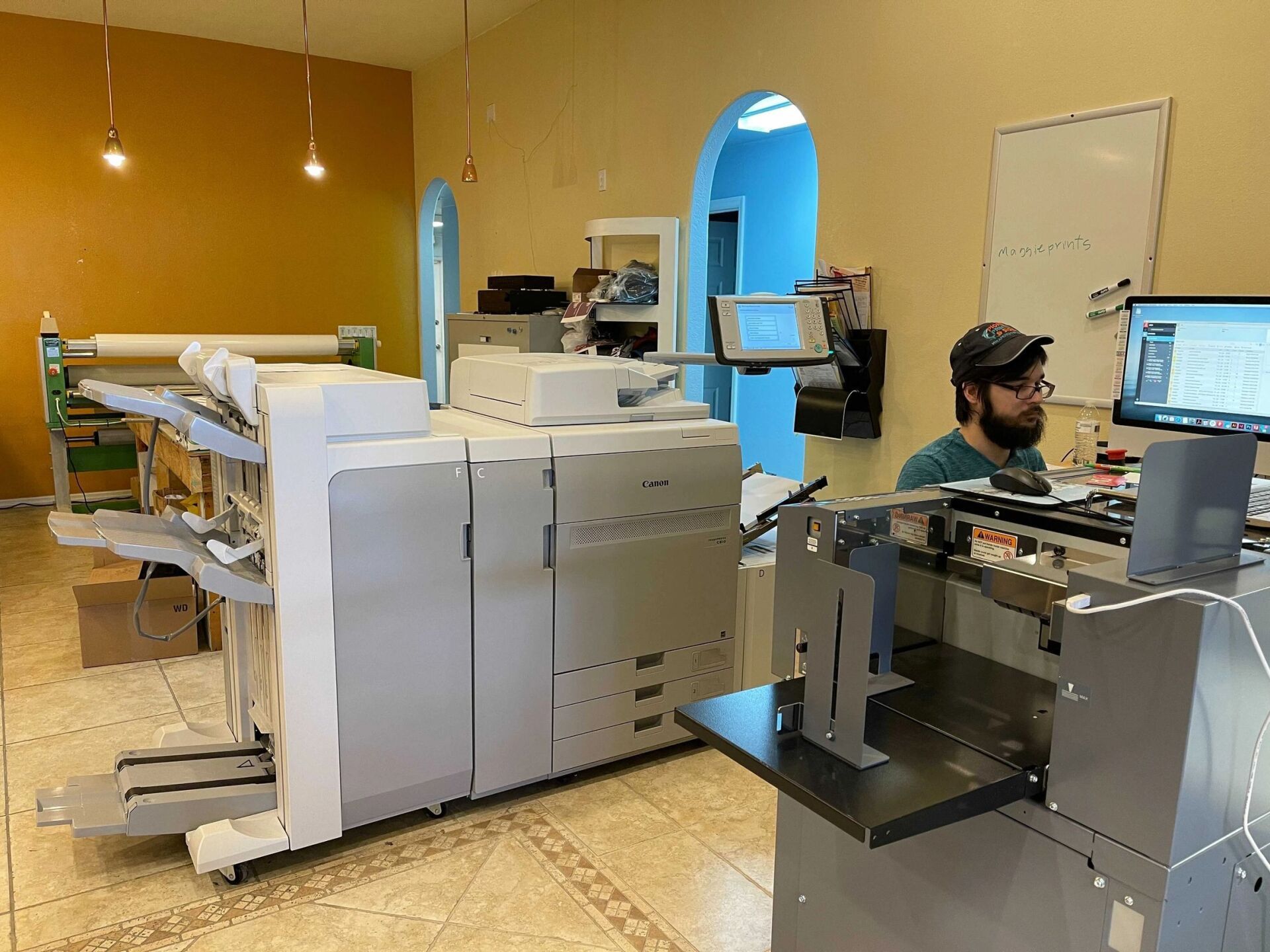 A man works at a computer in a print shop next to large printers and a cutting machine.