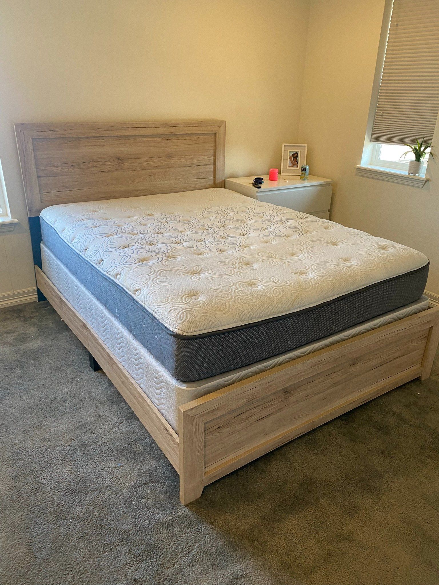 Wooden bed with mattress in a bedroom, near a nightstand and window.