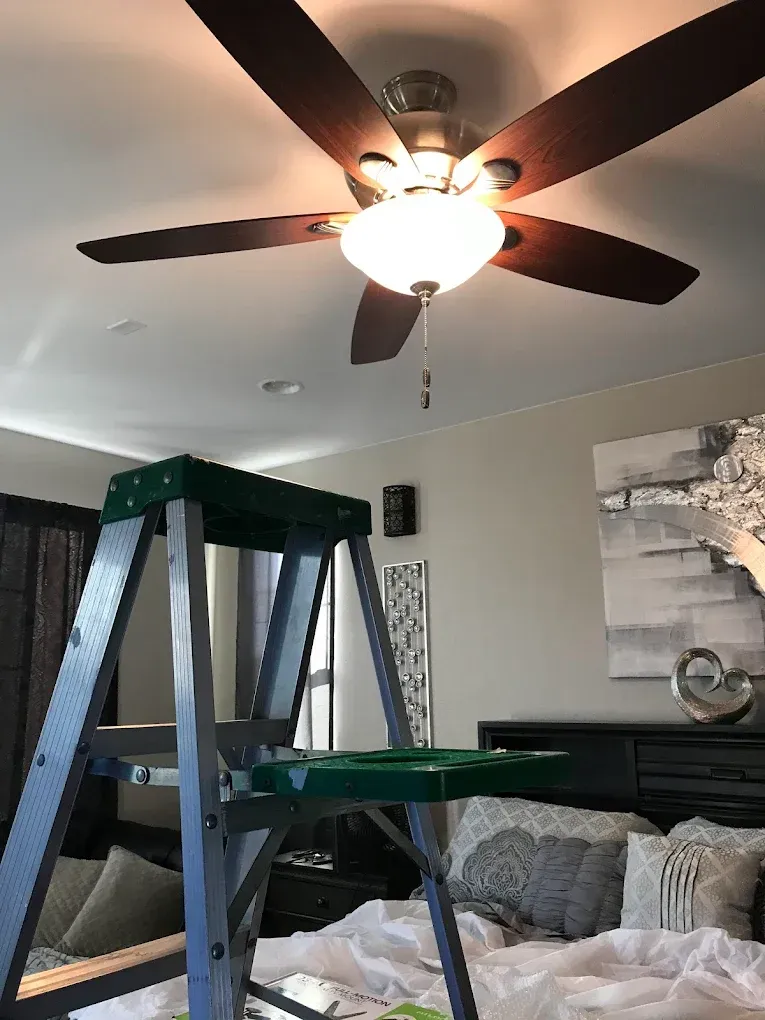 Ladder beneath a ceiling fan in a bedroom, likely for maintenance or repair.