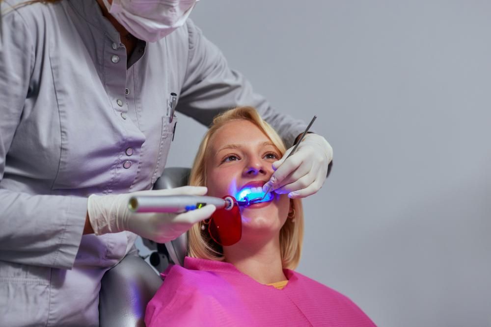 A woman is sitting in a dental chair getting her teeth whitened by a dentist.