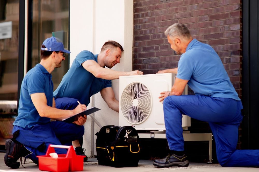 Three technicians installing an outdoor AC unit. One kneels, writing notes, while two others work on the unit.