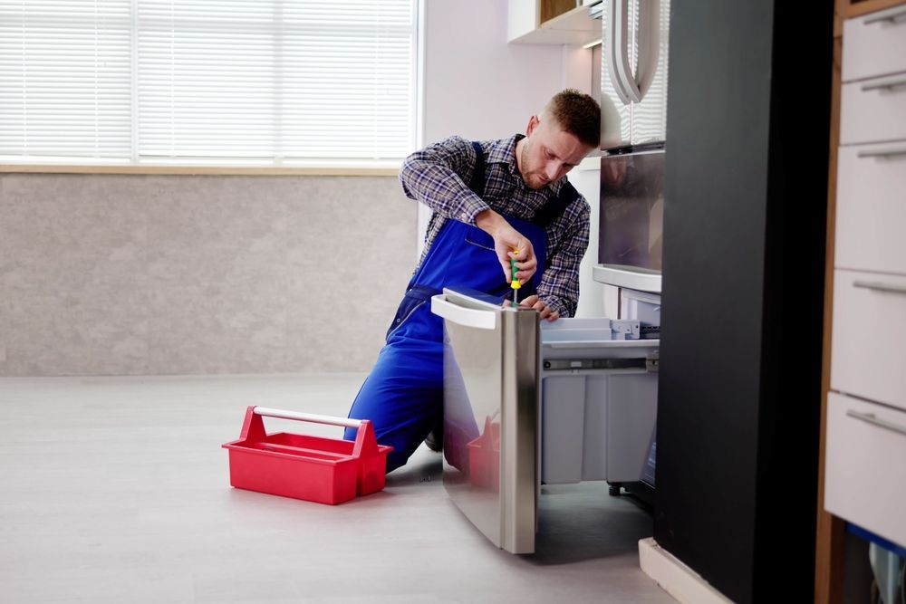 A repairman in blue overalls fixing a refrigerator with a screwdriver, red toolbox nearby.