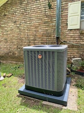 Gray air conditioning unit on a dark square base next to a brick wall and window.