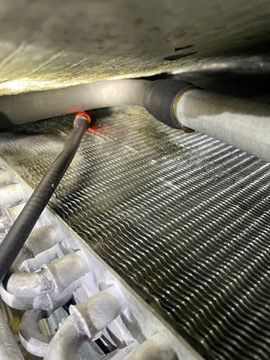 Tool probing an air conditioning unit's evaporator coils, near black hose and silver components.