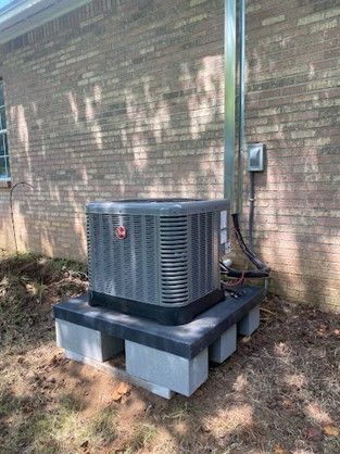 Air conditioning unit on a cinder block base, next to a brick building.