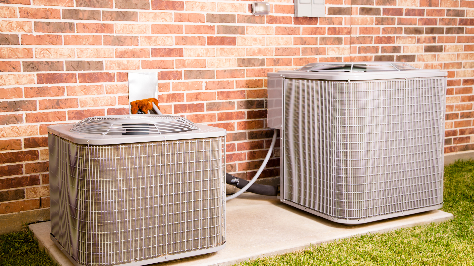 Two gray air conditioning units sitting on concrete pad against a brick wall.