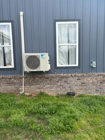 A Daikin mini-split AC unit on a blue-sided house with brick base and two windows. White piping and a white vertical exhaust pipe are also visible.
