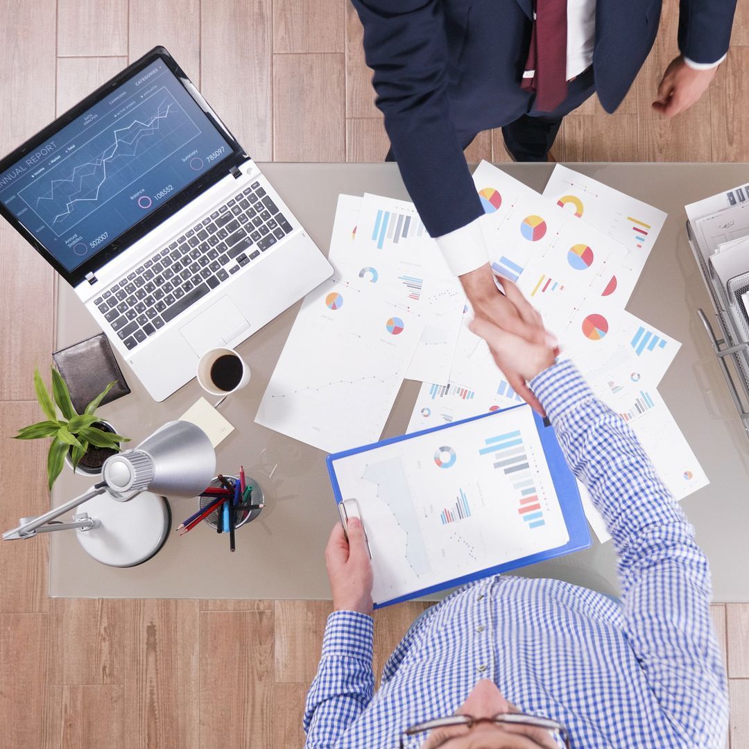 Two men shaking hands over a desk with papers and a laptop