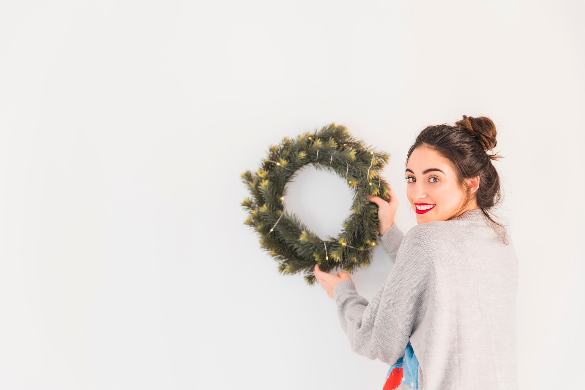 Woman hanging a Christmas wreath on a white wall, smiling with red lipstick.