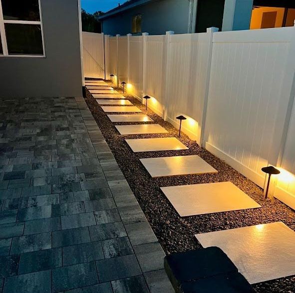 A brick walkway leading to a white fence with lights on it.