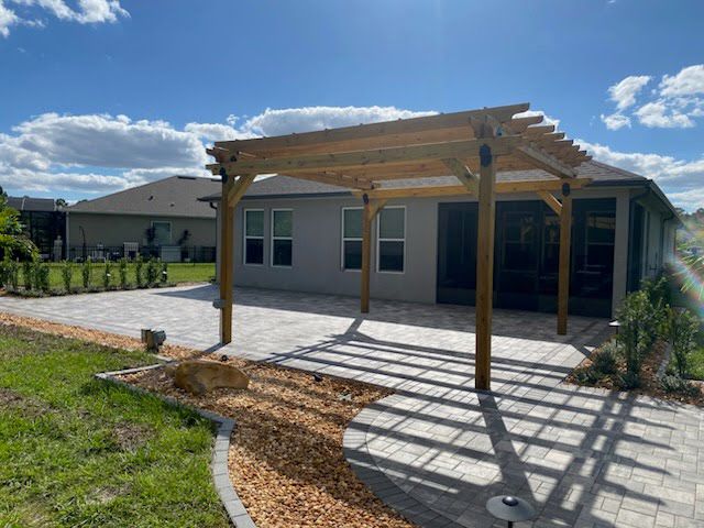 Pergola over a paved patio, extending from a house with a blue sky.