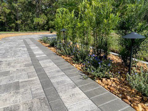 Paver driveway with landscaping, featuring lighting and gravel. Green plants and trees in background.