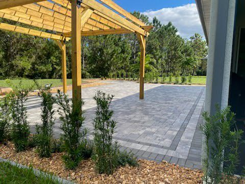 Wooden pergola over a gray paver patio with landscaping and house in the background.