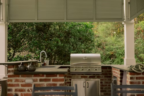 Outdoor kitchen with brick and stainless steel grill, sink, and green trees in the background.