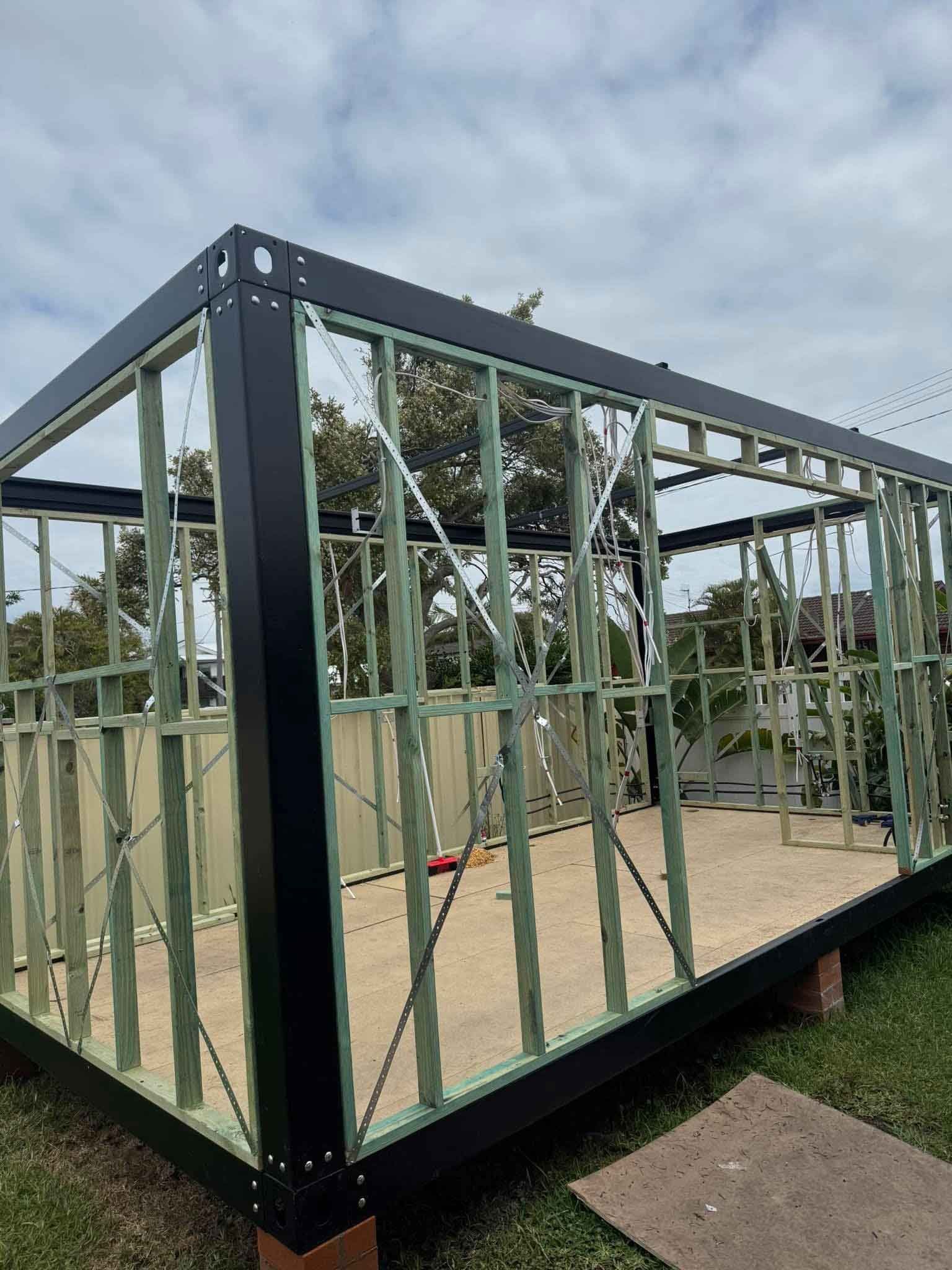Wooden frame structure under construction with black trim, against a cloudy sky.